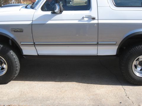 1994 Ford Bronco - The side profile of a white and grey 1994 Ford Bronco is visible, with a focus on the door and wheels.