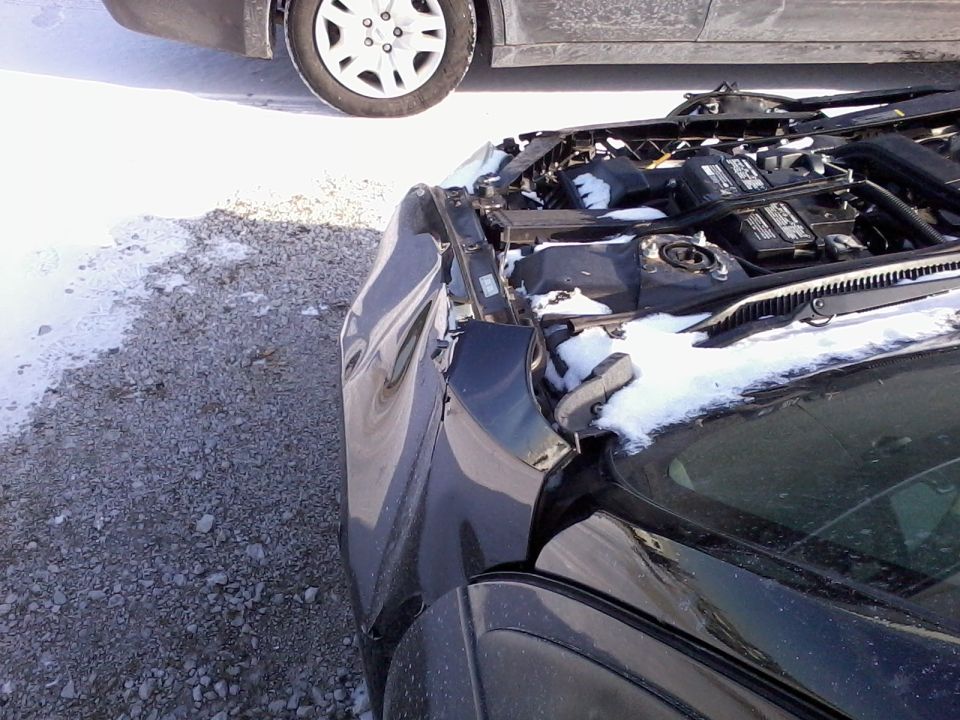Front end of a dark car with hood open and snow dusting the engine bay in a gravel lot