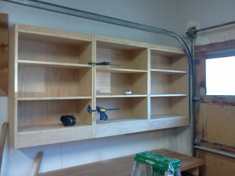 1996 Ford Bronco - A wooden shelving unit is mounted on a garage wall, with several clamps attached to the shelves and a black object on the lowest shelf.