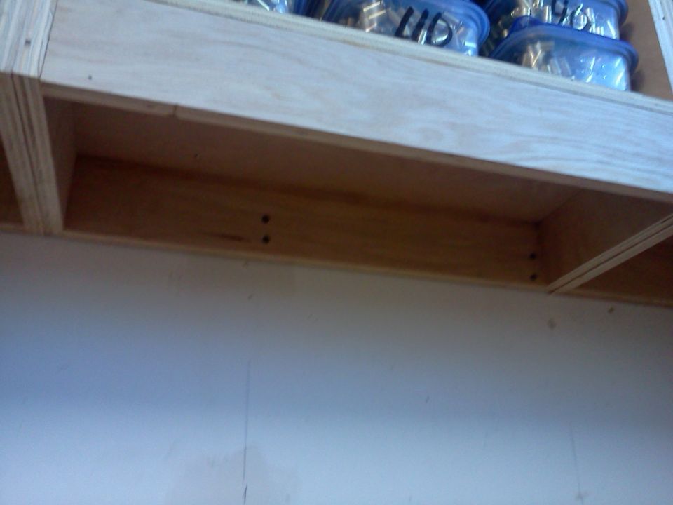 Wooden storage shelf holding clear plastic containers with items inside. Supermotors.net logo at bottom right