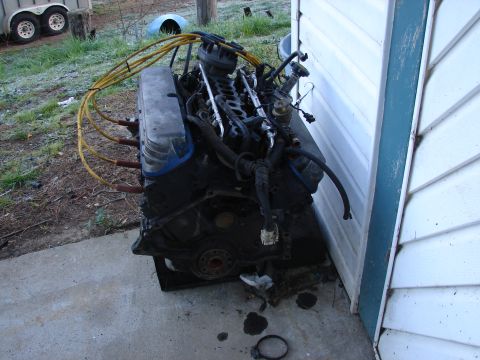 1973 Ford Ranchero - A V8 engine block with spark plug wires and other components is resting on the ground outdoors.