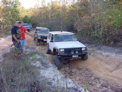 1986 Ford Bronco - A group of off-road vehicles, including a white Jeep Cherokee XJ, navigate a dusty trail through a wooded area.