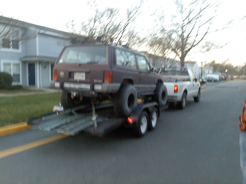 White Ford pickup towing a lifted maroon Jeep Cherokee on an open trailer along a residential street