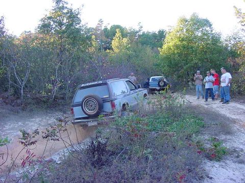 1986 Ford Bronco - A group of people observe off-road vehicles navigating a challenging dirt path in a wooded area.