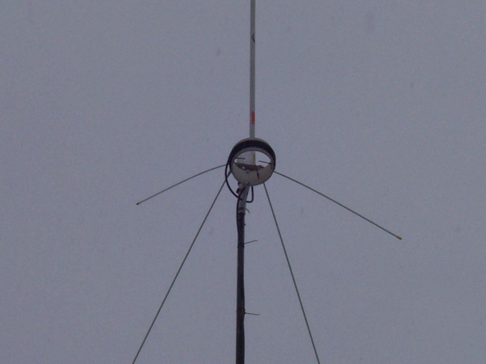 Close-up of a radio antenna with wires extending outwards against a gray sky. Supermotors.net logo visible