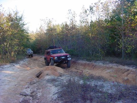 1986 Ford Bronco - Two off-road vehicles tackle a challenging trail in a wooded area.