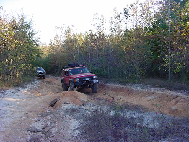 Red lifted Jeep Cherokee XJ with roof rack climbing out of a rutted clay trail rut