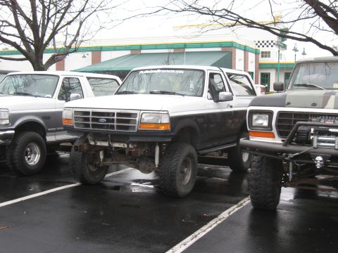 1994 Ford Bronco - Multiple lifted trucks are parked in a lot, with one white and black Ford Bronco prominently displayed.
