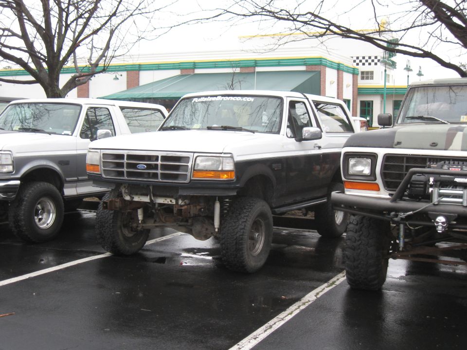 White and black Ford Bronco with lifted suspension and off-road tires, parked on wet asphalt