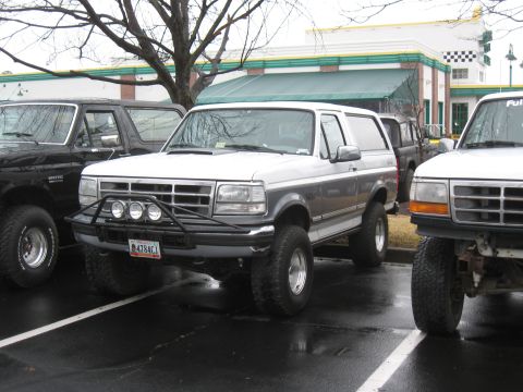 1994 Ford Bronco - A 1994 Ford Bronco is parked next to other trucks, a custom front bumper with auxiliary lights.