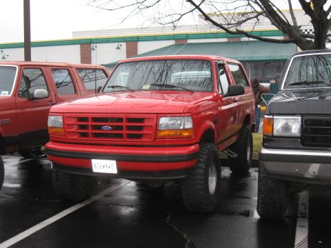1994 Ford Bronco - A red Ford Bronco is parked in a lot next to other vehicles on a cloudy day.
