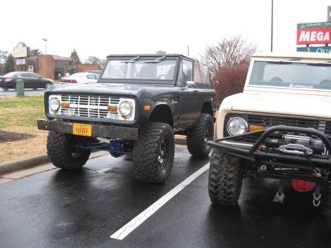 1994 Ford Bronco - Two Ford Broncos, one dark and one light, are parked on a wet asphalt surface.