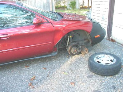 1997 Ford Taurus - A red 1997 Ford Taurus is missing its front passenger wheel, with the tire lying on the ground nearby.