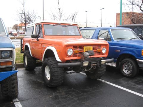 1994 Ford Bronco - An orange Ford Bronco with a white top and large off-road tires is parked in a lot next to other vehicles.