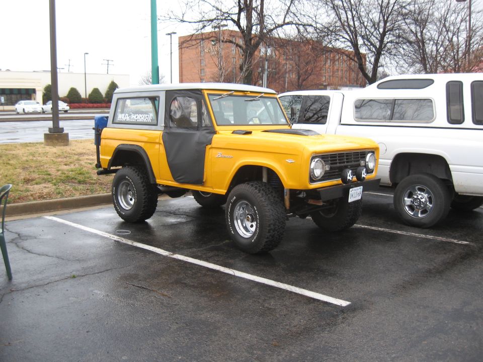 Yellow Ford Bronco with black soft doors parked next to a white pickup truck with a camper shell. License plate reads "SKOOBAR"