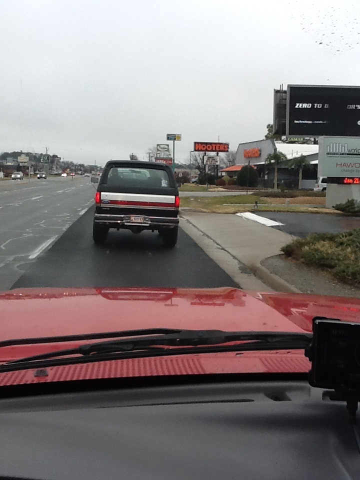 Red hood POV following a dark two-tone Ford Bronco on a wet road past a Hooters sign
