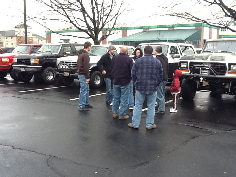 People gathered in a wet parking lot in front of several Ford trucks and Broncos, including a lifted gray Bronco with winch bumper