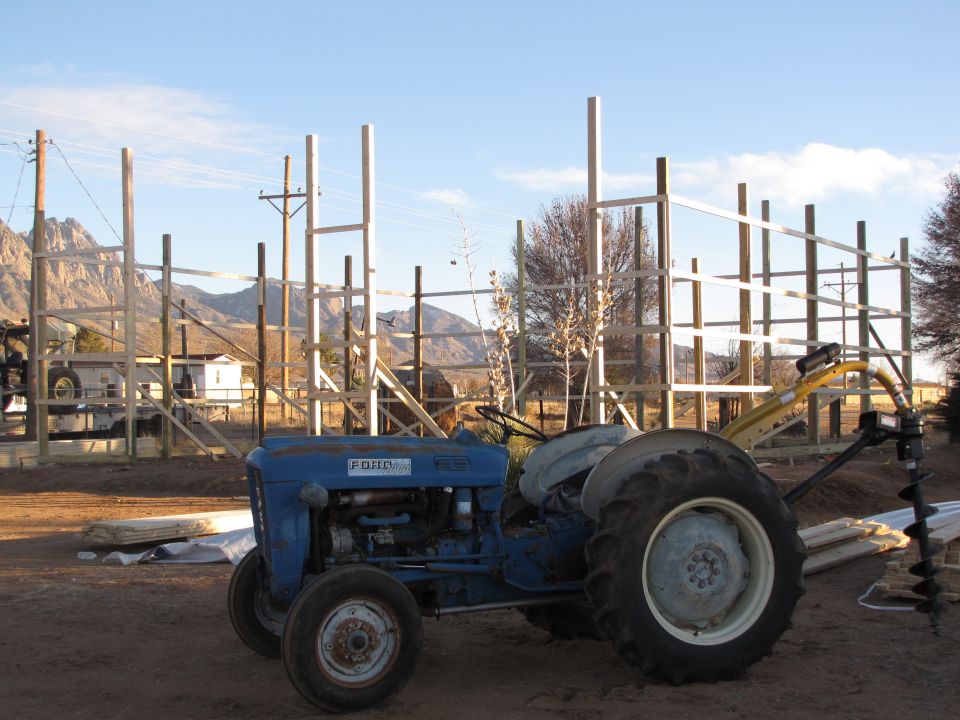 Blue Ford tractor with a yellow auger attachment, parked outdoors near a wooden structure under construction