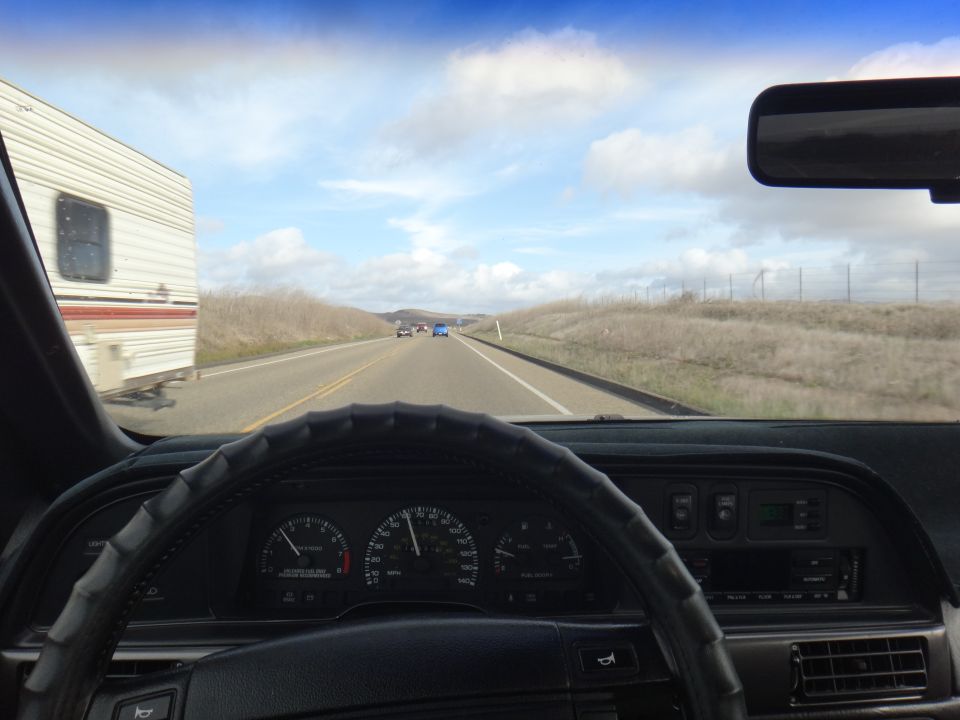 Dashboard and steering wheel of a car on a highway, with a trailer in the left lane and other cars ahead