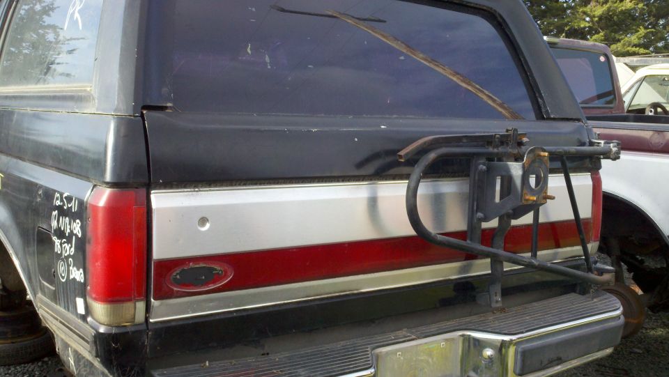 Rear of black Ford Bronco with red and silver stripe, spare tire carrier attached. Another vehicle is partially visible behind it