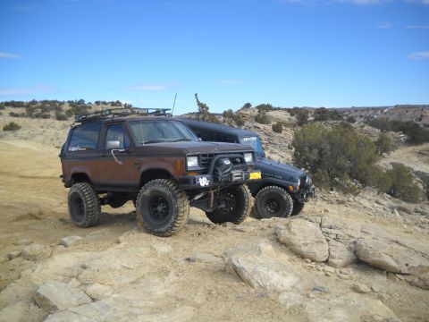 1992 Ford Explorer - Two off-road vehicles, a brown Ford Bronco II and a dark Jeep Wrangler, are parked on a rocky, desert-like landscape under a clear blue sky.