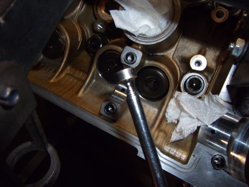 Close-up of engine cylinder head with valves and a socket wrench. Paper towels are visible. Supermotors.net logo at bottom