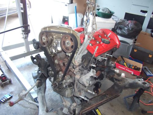 Engine assembly suspended by a chain hoist, with a red valve cover and timing belt visible