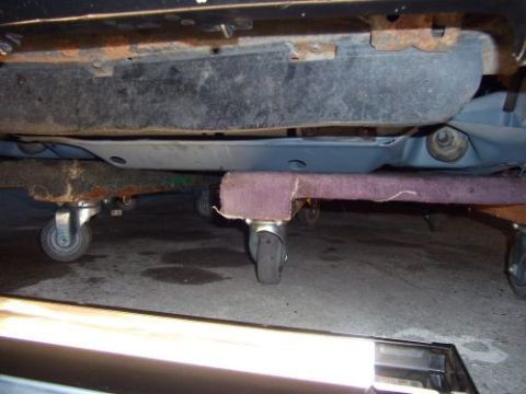 1991 Ford Taurus - The underside of a vehicle is shown, supported by a rolling creeper jack. The vehicle appears to be a sedan with visible rust on its frame.