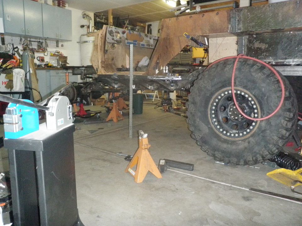 Vehicle frame and large tire on jack stands in a workshop. Metal fabrication tools are visible