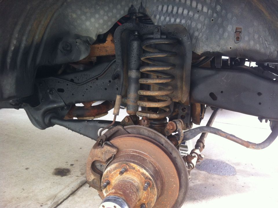 Rusty front suspension and brake assembly of a vehicle, showing coil spring, shock absorber, and brake rotor