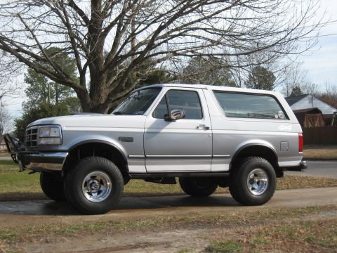 1994 Ford Bronco - This two-tone Ford Bronco is shown from the side, parked on a dirt road with a large tree and houses in the background.