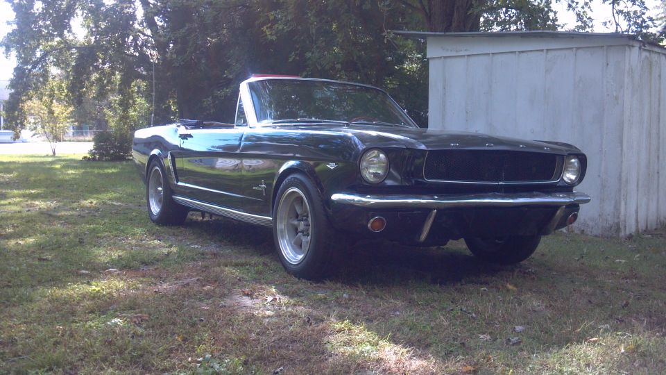Black 1965 Ford Mustang convertible parked on grass near a white shed