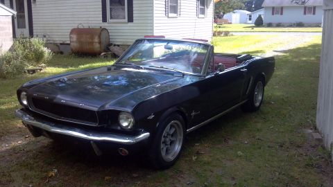 1965 Ford Mustang - A black 1965 Ford Mustang convertible with a red interior sits in a grassy yard next to a house.