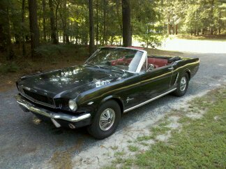 1965 Ford Mustang - A classic black 1965 Ford Mustang convertible sits parked on a gravel road surrounded by trees.