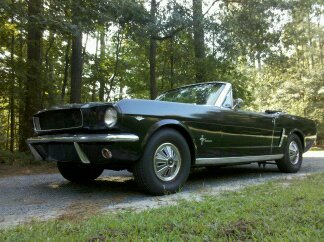 1965 Ford Mustang - A black 1965 Ford Mustang convertible is seen from a 3/4 front angle on a dirt road surrounded by trees.