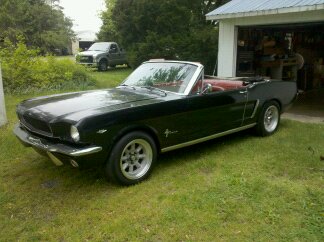 1965 Ford Mustang - A black 1965 Ford Mustang convertible is parked on the grass next to a garage, with a pickup truck in the background.