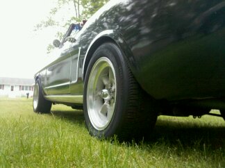 1965 Ford Mustang - A dark 1965 Ford Mustang convertible is parked on a grassy lawn, showing its rear wheel and quarter panel.