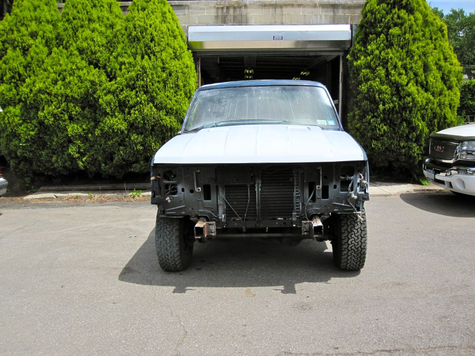 Front view of a 1992 Ford Bronco with its bumper and grille removed, parked outside a garage. Another truck is visible to the right