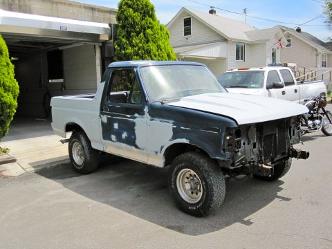 1992 Ford Bronco - A Ford Bronco is parked outside a garage, showing signs of repair work and missing front-end components.