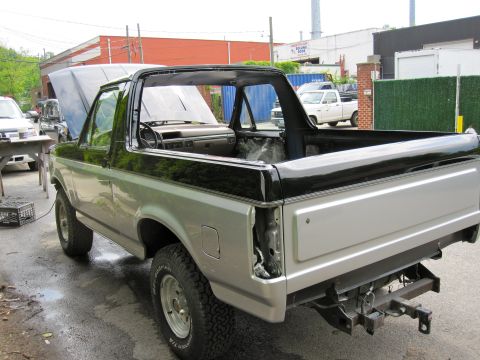 1992 Ford Bronco - The rear passenger side of a silver and black Ford Bronco is shown with the hood and roof removed. The vehicle is parked on a wet asphalt s…