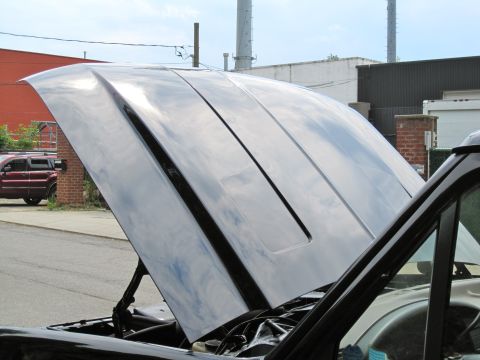 1992 Ford Bronco - The glossy black hood of a vehicle is open, revealing the engine bay. In the background, a red SUV is parked near a brick building.
