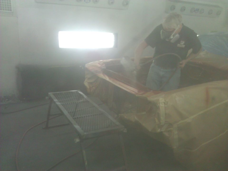 Man in respirator sprays a car body covered in plastic sheeting inside a spray booth