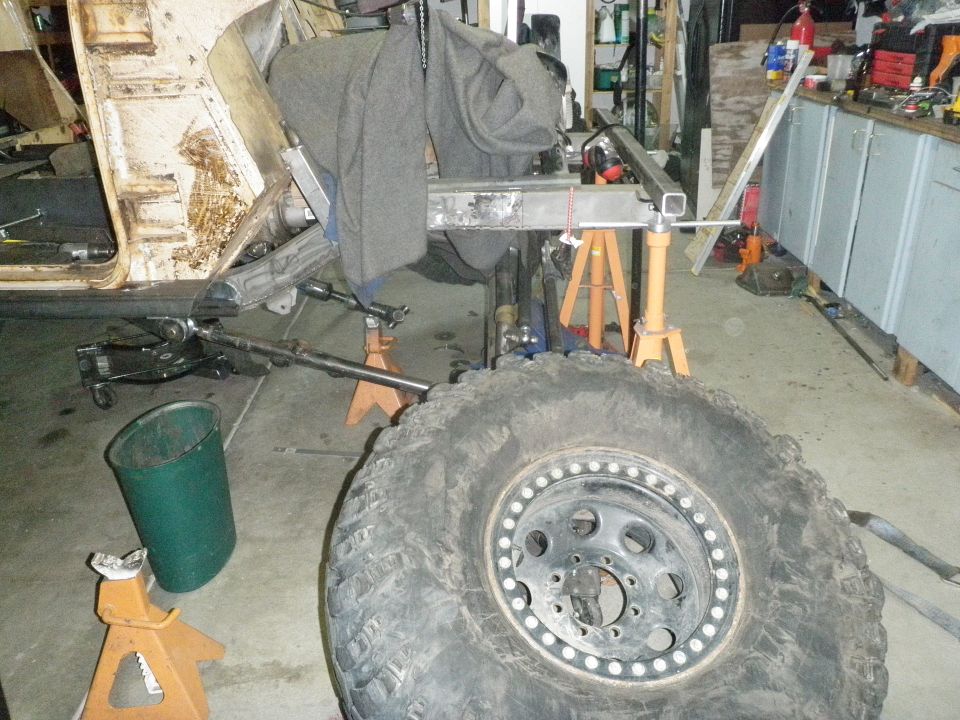 Large off-road tire and wheel in front of a disassembled vehicle chassis on jack stands.