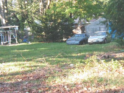 1997 Ford Taurus - Two light-colored sedans parked side by side on a grassy back lot beside a weathered gray shed and a swing set, framed by tall trees and a…