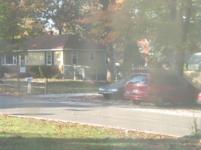 1997 Ford Taurus - A hazy, backlit shot across a residential street of a red Ford Taurus sedan parked at the curb nose-to-tail with a dark gray sedan, in fron…