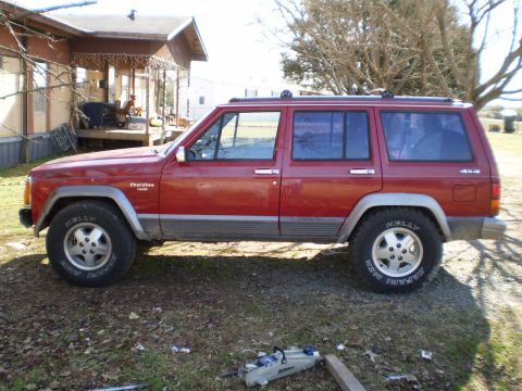 1992 Jeep Cherokee - A red Jeep Cherokee is parked on a grassy area next to a house, with a "Cherokee" badge visible on the fender.