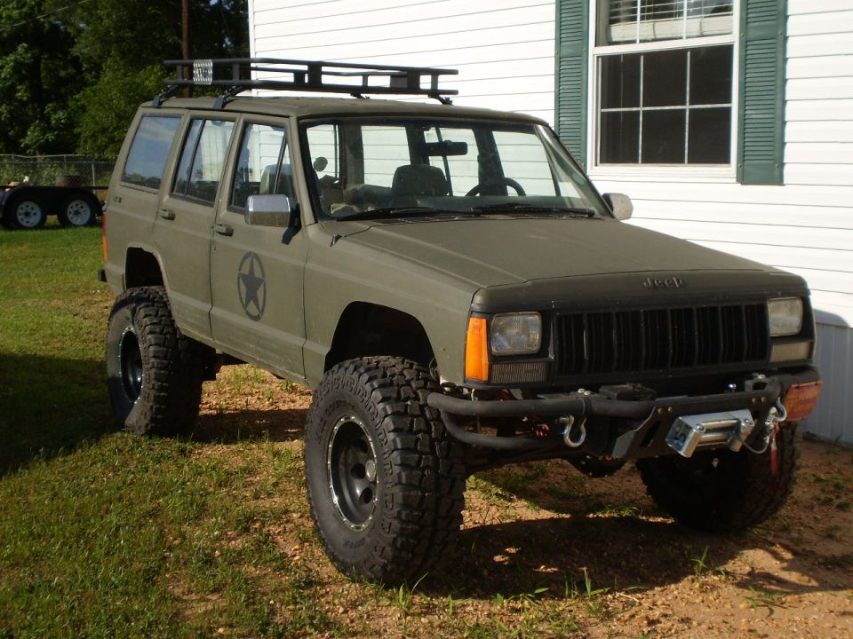 Olive drab Jeep Cherokee with a roof rack, winch, and large off-road tires parked in front of a white house