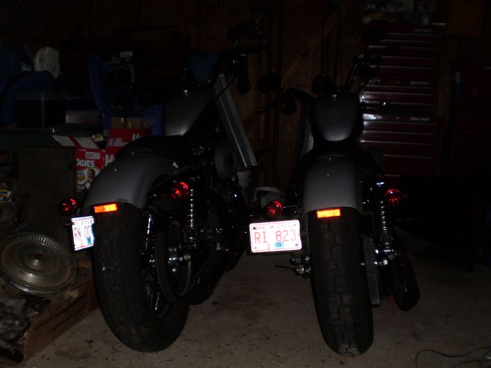 Two dark motorcycles parked in a garage, rear view. One has license plate RI 823