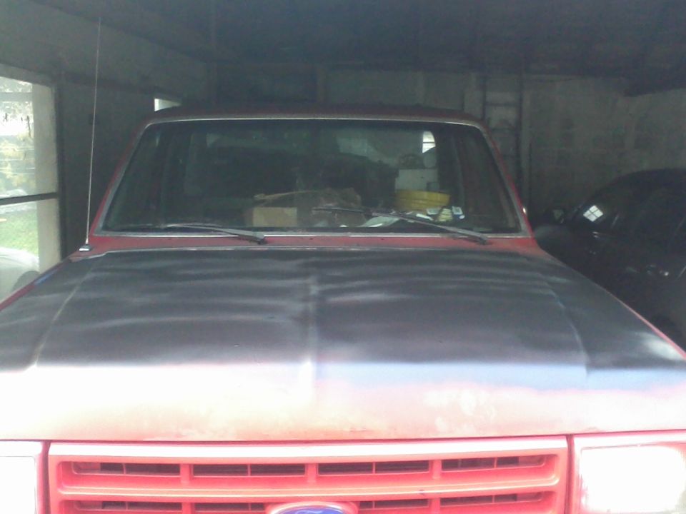 Red truck's front end, hood is unpainted. Another dark vehicle is parked behind it