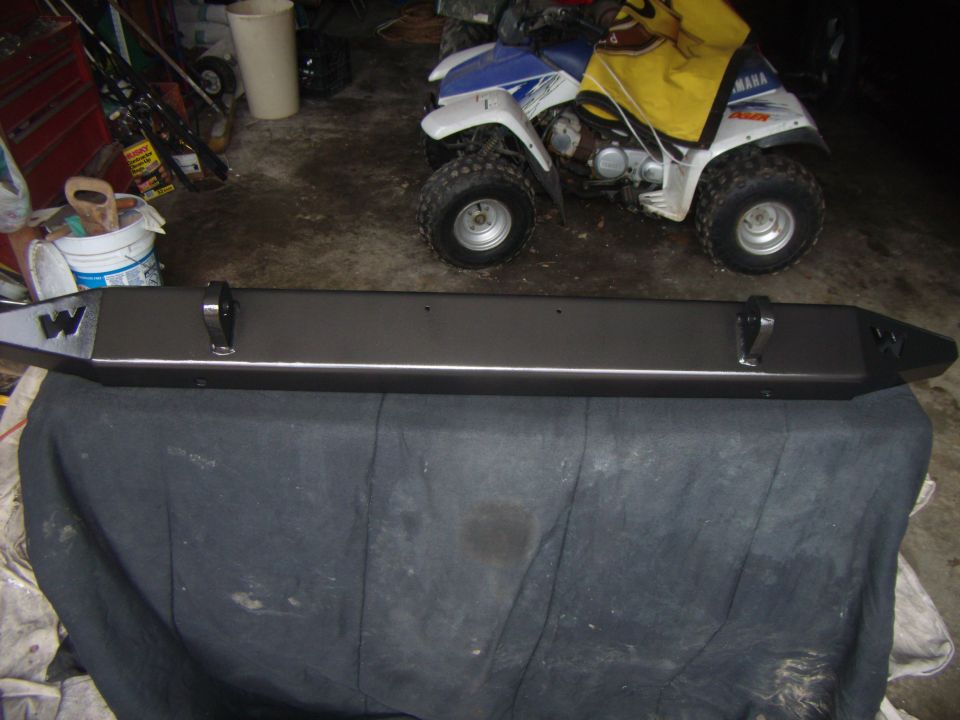 Aftermarket bumper with tow hooks and a 'W' logo on the left end, resting on a dark cloth